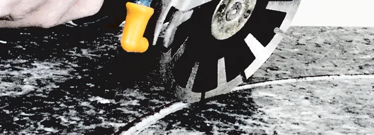 A close-up of a circular saw with a yellow handle cutting through a slab of grey granite. The blade is in motion, creating a curved cut in the stone.