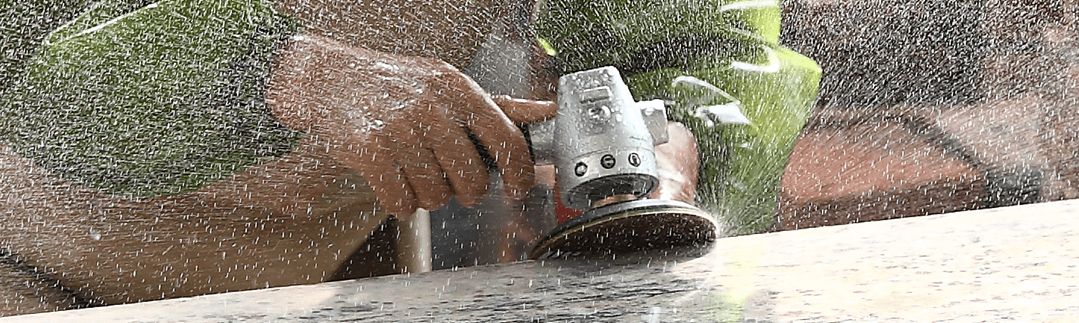 A person in work attire polishes a stone surface using a power tool, creating a spray of water and fine particles. The close-up captures the motion and energy of the task.