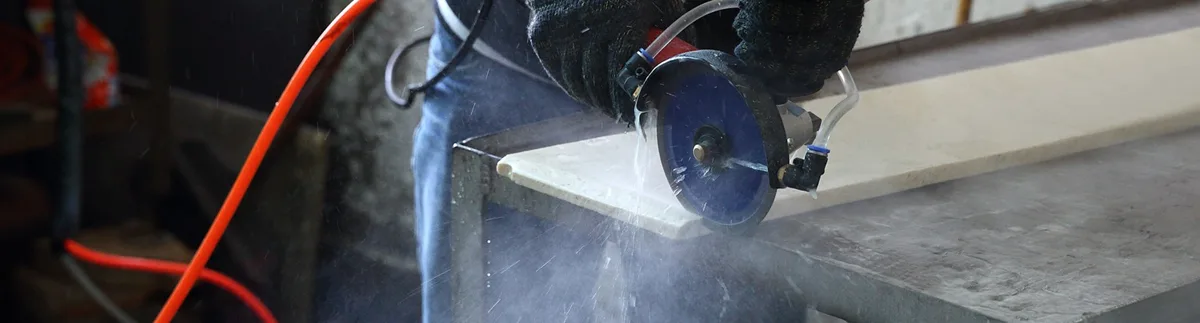 A person wearing gloves uses a power saw to cut a wooden plank, creating sawdust and debris. The scene shows a close-up of the saw in action, with a red hose visible in the background of the workshop.