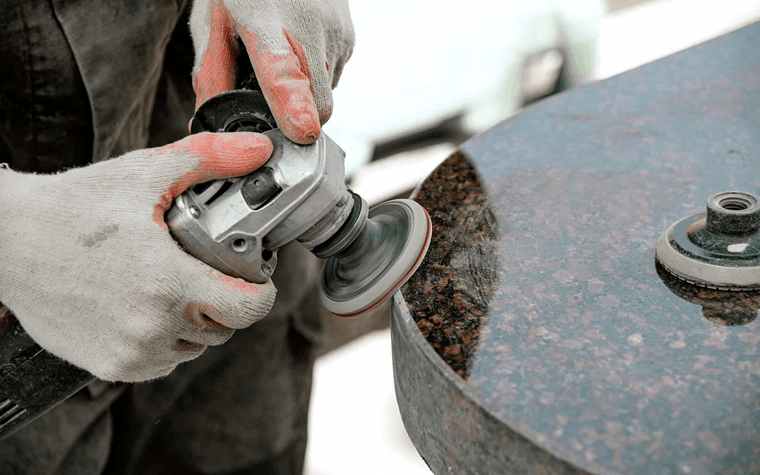 A person wearing white gloves uses a handheld power tool to polish the edge of a granite countertop, with visible motion blur on the tool indicating movement. The surface has a glossy finish.