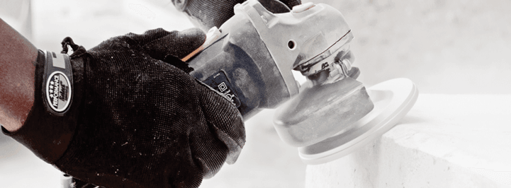 A person wearing black gloves uses a handheld power tool to polish or sand a white surface. The tool is gray and blue, and fine dust is visible, indicating active work.