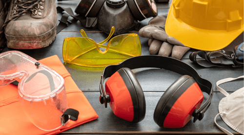 A collection of safety equipment including earmuffs, a hard hat, goggles, safety glasses, respirators, work gloves, a high-visibility vest, and work boots is displayed on a dark wooden surface.